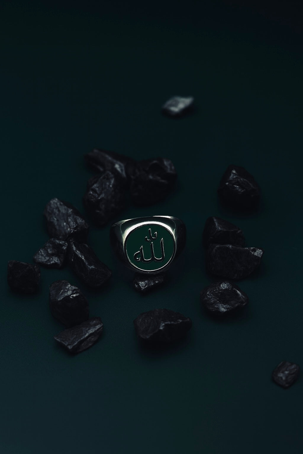 Mutawakkil Hikmah green Islamic ring engraved with Allah in Arabic calligraphy, placed among dark stones on green surface.
