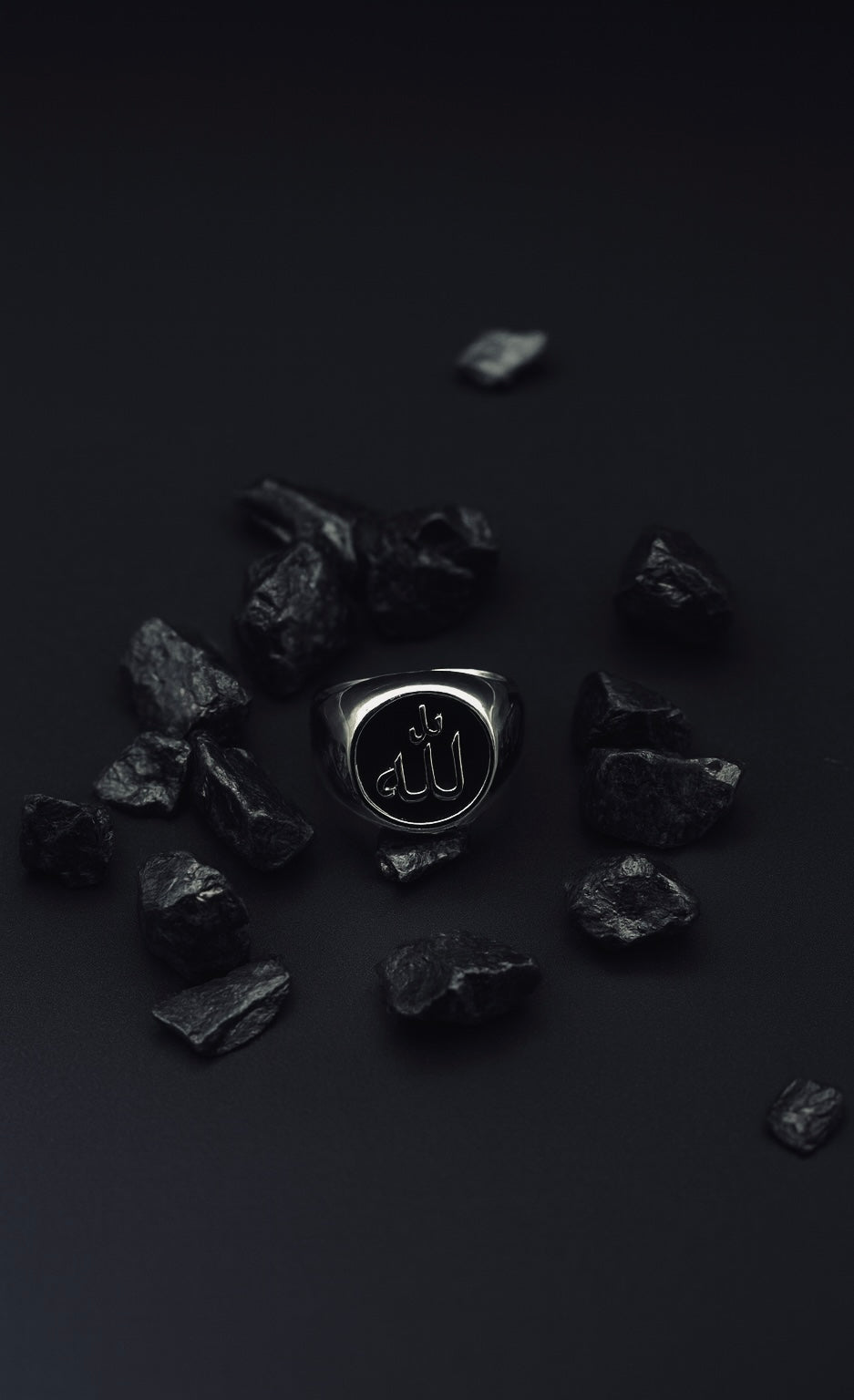 Mutawakkil Quwwah black Islamic ring engraved with Allah in Arabic calligraphy, placed among dark stones on black surface.