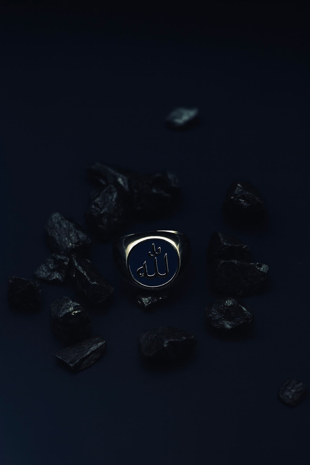 Mutawakkil Sabr blue Islamic ring engraved with Allah in Arabic calligraphy, placed among dark stones on deep blue surface.