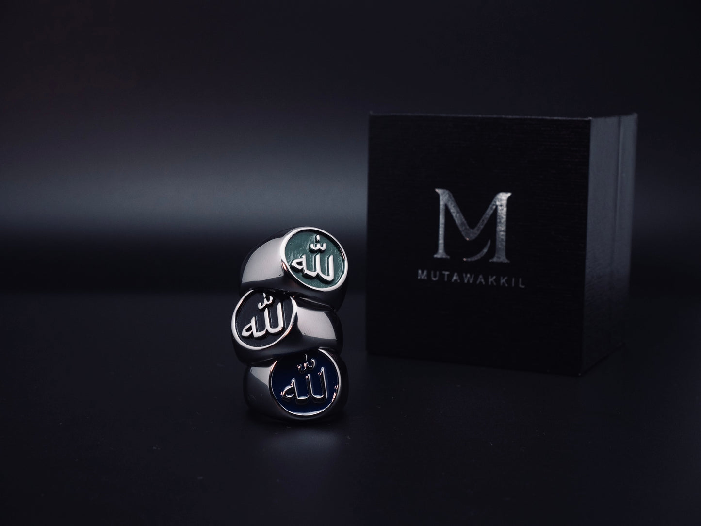 Set of Mutawakkil Islamic rings engraved with Allah in Arabic calligraphy, featuring black, blue and green models beside luxury box.
